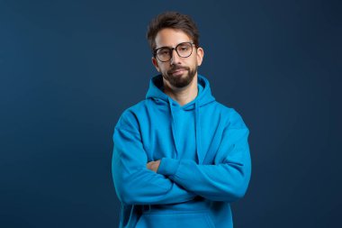 Man wearing glasses and blue hoodie looking disinterested, standing with arms crossed and slightly downcast gaze, embodying sense of boredom or disappointment, posing against dark background clipart
