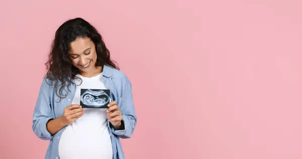 年轻快乐的黑发孕妇 腹部丰满 展示婴儿的超声图像 胸部上方挂着胎儿照片 有粉色工作室背景 全景与复印空间 — 图库照片