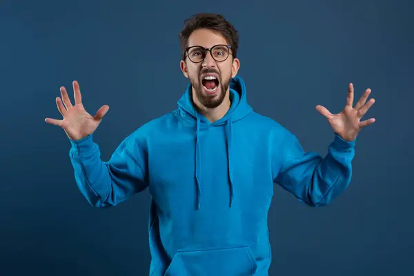 Stock image Portrait Of Angry Young Man Screaming At Camera With Rage, Furious Aggressive Guy Wearing Eyeglasses Raising Hands And Emotionally Shouting, Standing Against Blue Background In Studio