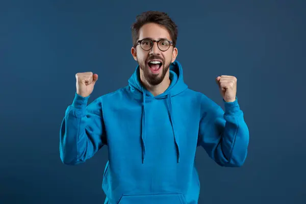 stock image Joy Of Win. Emotional Young Man Celebrating Success With Raised Fists, Overjoyed Millennial Guy Wearing Blue Hoodie Cheering Victory, Standing Against Blue Studio Background, Free Space