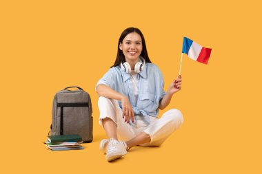 Neşeli kız öğrenci gururla Fransız bayrağı sergiliyor, küresel eğitim ve kültür alışverişi ile olan bağlantısını simgeliyor, teknoloji meraklısı akademik yaşam tarzı