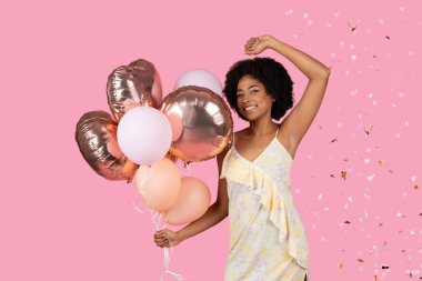 Neşeli Afrika kökenli Amerikalı kadın neşeyle dans ediyor, elinde gül sarısı ve pembe balonlar, etrafında konfetiler uçuşuyor, neşeli pembe bir arka plan, stüdyo