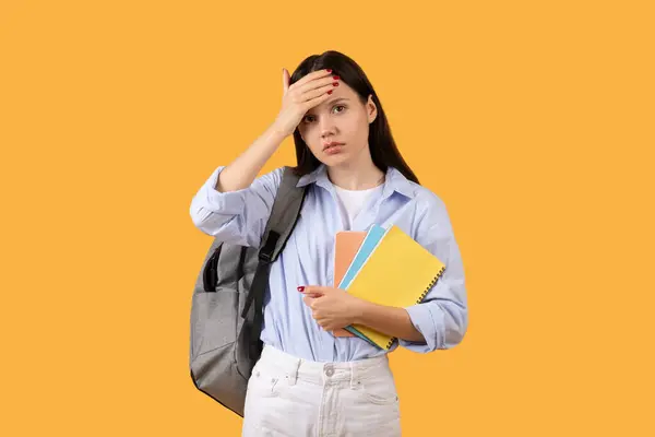 忧心忡忡的女学生背着背包和彩色笔记本站在黄色的背景下 用焦虑 压力或困惑的姿势触摸着额头 — 图库照片