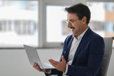 Mavi takım elbiseli orta yaşlı Avrupalı işadamı bilgisayarına bakarken gülümsüyor ve el kol hareketi yapıyor. Potansiyel olarak sanal bir toplantıda ya da Webinar 'da sunum yapıyor. İş, iş.