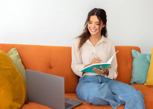 Young Smiling Beautiful Woman Taking Notes While Study With Laptop At Home, Happy Female Noting Down Information From Computer While Sitting On Couch In Living Room, Enjoying Online Education