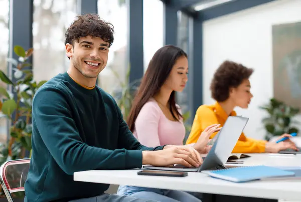 Smiling male student with curly hair sitting with diverse female classmates in sunny, plant-filled study space, all focused on their individual laptops