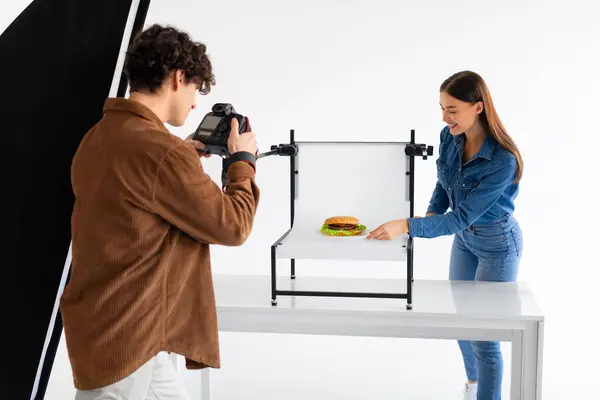 Skilled food photographer in brown jacket intensely focuses his camera on perfectly styled burger, with an assistant adjusting the presentation on white tabletop