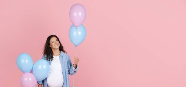 Kocaman göbekli, pembe ve mavi balonları olan genç ve güzel bir kadın. Cinsiyet kutlaması yapıyor. Pembe arka planda izole edilmiş.