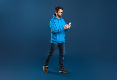 Mavi Stüdyo Arkaplanı Üzerinde Akıllı Telefonla Yürüyen Genç Tarz Adamı, Arkadaşlarıyla Cep Telefonu ya da Alışveriş İnternetindeki Yakışıklı Milenyum Adamı Mesajlaşması, Kopya Alanlı Tam Uzunluk