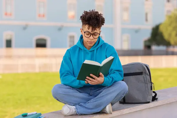 Pensive black student guy sitting with book and backpack, reading outdoors at university campus park, learning literature. Bookworm wearing eyeglasses reads new novel outside