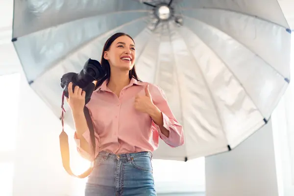 一个快乐的年轻女人拿着相机 自信地把大拇指放在一个大工作室的轻伞下 穿着粉色衬衫和斜纹棉布牛仔裤 自由自在 — 图库照片