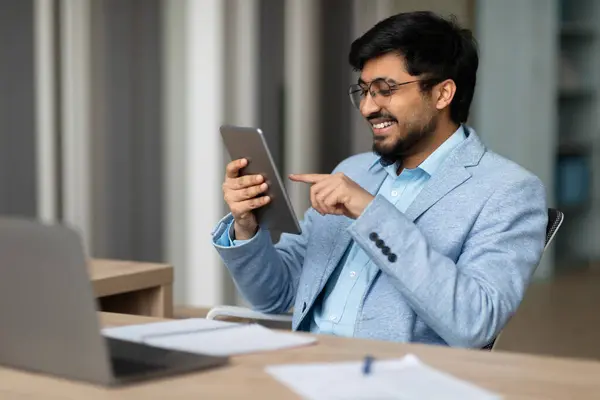 Happy Indian businessman at modern office desk with laptop browsing on his tablet indoors, browsing and communicating online. Corporate lifestyle and modern approach to work and technology