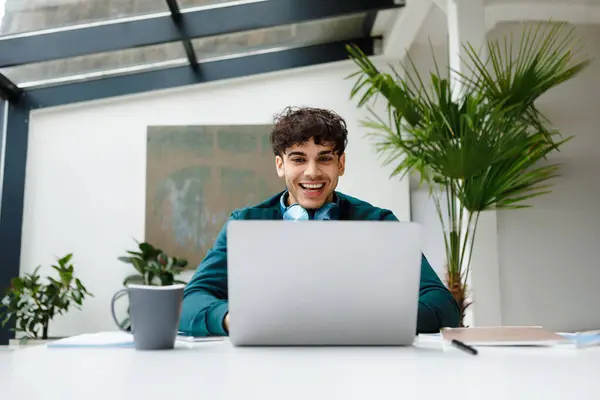 Joyful young freelancer man with trendy headphones laughs while engaging with content on his laptop in sunny, plant-filled workspace, showing enjoyment