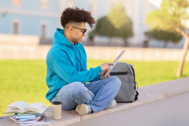 Siyahi öğrenci dijital tabletle geziniyor ve öğreniyor, paket kahve ve çalışma kitaplarıyla birlikte açık kampüs parkında oturuyor, internetten ders çalışıyor. E-öğrenim, modern eğitim