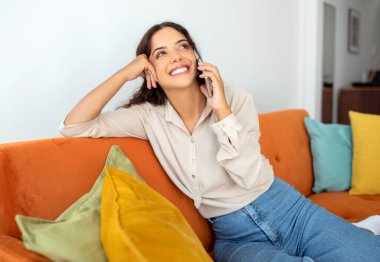 Mutlu genç kadın, günlük kıyafetiyle akıllı telefonunda keyifli sohbetler yapıyor, neşeli, güzel bayan kanepede uzanmış, parlak yastıklarla konuşuyor, cep telefonuyla konuşuyor, kapatıyor.
