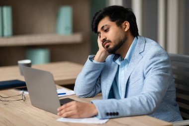 Sıkıcı iş. Sıkıcı Hintli iş adamı dizüstü bilgisayarda çalışırken başını dinlendirirken modern ofiste uykulu ve yorgun hissediyordu. İlgisiz Çalışan, Aşırı Çalışma ve İşyeri Yorgunluğu Konsepti