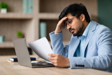 Gözlüklü stresli Hindu işadamı, iş gazetelerini okurken, bilgisayarında modern ofis içi bilgisayarını incelerken sorun yaşıyor. Evrak işlerinde endişeli girişimcinin yan görüntüsü