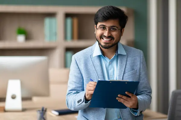 Professional Indian HR manager guy holds clipboard with signed contract, confident and successful entrepreneur signing completing business deal at modern workplace. Career offers, achievements