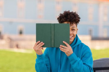 Kitap kurdu. Neşeli Afrikalı Amerikalı öğrenci, kameraya bakıyor kitap ile yüzünü kapatıyor, üniversite binasının dışındaki derslere hazırlanıyor. Edebiyat ve Eğitim