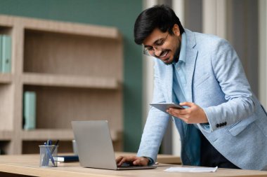 Gülümseyen Orta Doğulu işadamı cep telefonuyla mesajlaşıyor ve dizüstü bilgisayar kullanıyor, modern ofis içi iş ve profesyonel iletişimi yönetiyor, kapalı masa başında duruyor.