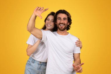 Neşeli Avrupalı genç çift kameraya doğru şakacı bir şekilde hareket ediyor, kadın elini kaldırıyor sanki bir anı yakalıyormuş gibi, ve adam işaret ediyor, hepsi de parlak sarı bir zemin üzerinde
