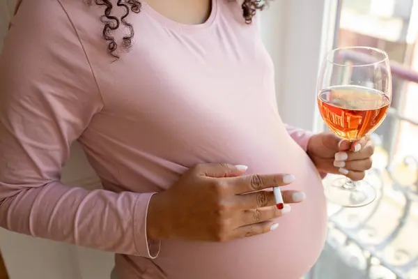妊娠中の悪い習慣について 自宅の窓の近くに立っている間 タバコとワインのグラスを保持している妊娠中の女性 禁煙された喫煙と飲酒中毒の認知できない妊婦 — ストック写真