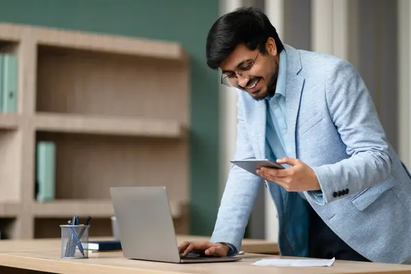Smiling Middle Eastern businessman texting on cellphone and using laptop, managing work and professional communication at modern office interior, standing near desk indoor