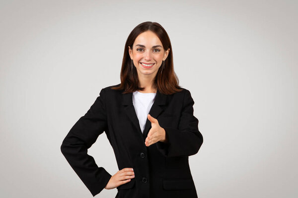 Approachable and professional caucasian young businesswoman with long hair wearing a black suit extends a handshake, suggesting a welcoming gesture or agreement, isolated on gray background, studio