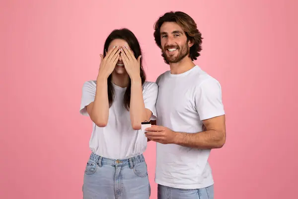 令人惊讶的是 欧洲女人用手捂住眼睛 笑容满面的男人拿着一个小珠宝盒 可能会向工作室求婚 两人都穿着粉色背景的白色T恤衫 — 图库照片