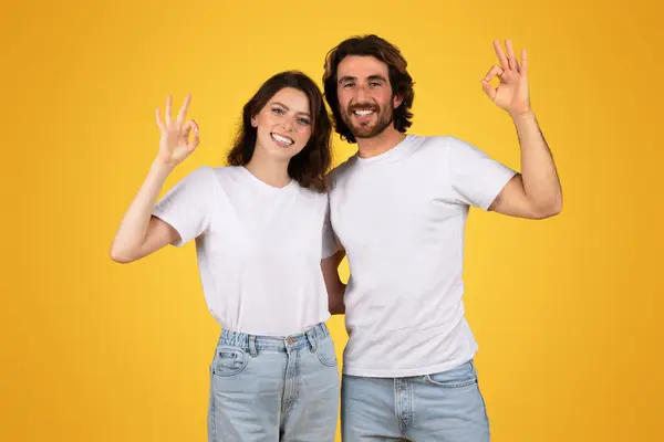 穿着白色T恤衫和牛仔裤的快乐的欧洲男人和女人做着一个确定的手势 带着温暖的黄色背景自信地微笑 象征着满足感和积极向上 — 图库照片