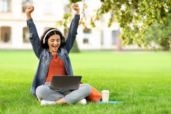 Excited young eastern woman student celebrating success, checking test results online, passed exam. Happy indian lady sitting on lawn in university campus, using laptop and wireless headphones