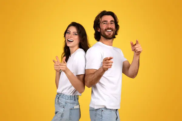 年轻的欧洲女人和留胡子的男人背靠背地站着 拿手指开玩笑地指着相机 穿着休闲的白色T恤衫和斜纹棉布 背景是明亮的黄色 — 图库照片