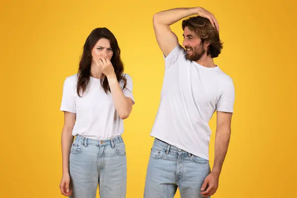 一个欧洲女人带着厌恶的表情捏着鼻子 而一个男人笑着挠了挠他的头 两个人都穿着白色的T恤衫和牛仔裤 背景是赤黄的 — 图库照片