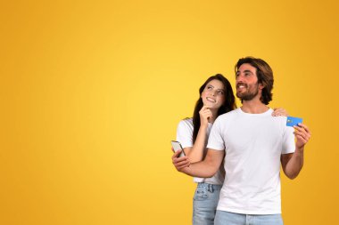 Avrupalı bir kadın rüyasında yukarı bakıyor ve elinde kredi kartı ve telefon olan sakallı bir adam gülümsüyor. Beyaz tişörtlü, sarı bir arka planda, stüdyoda alışveriş yapmayı planlıyor.