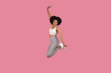 Kıvırcık saçlı, beyaz spor sutyen ve gri tayt giymiş, pembe arka planda soyutlanmış neşeli bir şekilde havada zıplayan enerjik genç Afro-Amerikalı kadın.