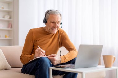 Evdeki web sitesine katılan heyecanlı yaşlı adam, koltukta oturuyor, dizüstü bilgisayar ekranına bakıyor ve not defterine notlar alıyor, kulaklık takıyor, boşluğu kopyalıyor. Yaşlılar için E-eğitim
