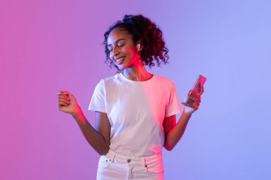 Kıvırcık saçlı, beyaz tişörtlü ve kulaklıklı neşeli genç bir kadın canlı pembe ve mavi arka planda akıllı telefon tutarken dans ediyor.