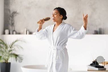Banyoda şarkı söylerken elinde mikrofon gibi saç fırçası tutan mutlu siyah kadın, beyaz bornozlu neşeli Afro-Amerikan kadın sabah hazırlanırken eğleniyor, bedava uzay