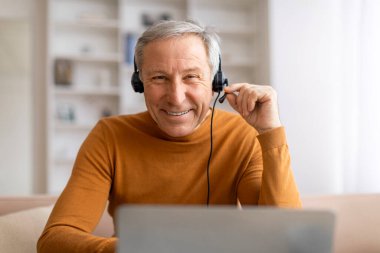 Pozitif dedemin dizüstü bilgisayar ve kulaklık kullanarak koltukta oturduğu yakın plan. Evden çalışan kıdemli bir adam, online görüşme, iş arkadaşlarıyla görüntülü konuşma. Yaşlılar için uzak iş