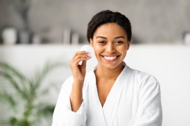 Pamuklu ped tutan neşeli siyah kadın, evde cilt bakımına hazır, beyaz bornozlu çekici Afro-Amerikan kadın, sakin banyo ortamında poz veriyor, kameraya bakıyor, uzay kopyası yapıyor.