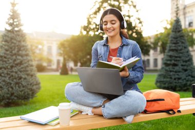 Çevrimiçi eğitim, uzaktan öğrenme konsepti. Genç Hintli kız öğrenci öğretmeniyle video görüşmesi yapıyor, Webinar 'a katılıyor, parkta bankta oturuyor, dizüstü bilgisayar ve kulaklık kullanıyor, notlar alıyor, işaret fişeği atıyor.