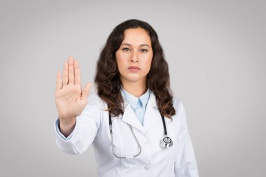 Beyaz önlüklü ciddi genç Avrupalı kadın doktor dur işareti yapıyor ve gri arka planda, stüdyoda izole edilmiş kameraya bakıyor. Hastalıklara karşı savaş