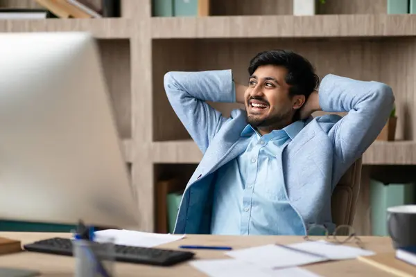 stock image Successful indian business guy resting holding hands behind head, relaxing in comfortable office chair at workplace after online work, sitting at table with desktop computer indoors