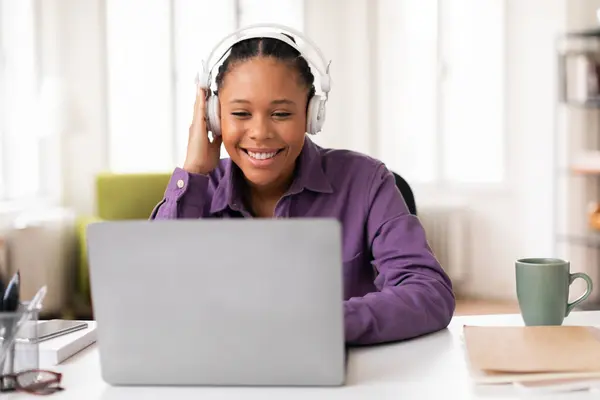 陽気な黒人女性学生がヘッドフォンを着て笑顔でラップトップスクリーンを見ながら 近くのノートブックとカップで勉強 — ストック写真