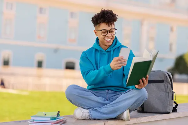 笑顔のブラジルの学生は 電子学習のためにスマートフォンを介してブックの写真を撮り 屋外の大学のキャンパスで彼の本やバックパックと座っている間 オンラインで勉強します フリースペース — ストック写真