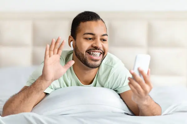 年轻快乐的黑人小伙子躺在床上 在智能手机屏幕前挥手微笑 用无线耳机 非洲裔美国千禧年男子与他的朋友们有视频通话 — 图库照片