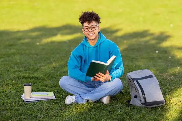 Educational picnic. Cheerful black student guy in eyeglasses reading book seated on green grass, learning with coffee outdoor at park, smiling to camera. Learner studying on lawn on weekend