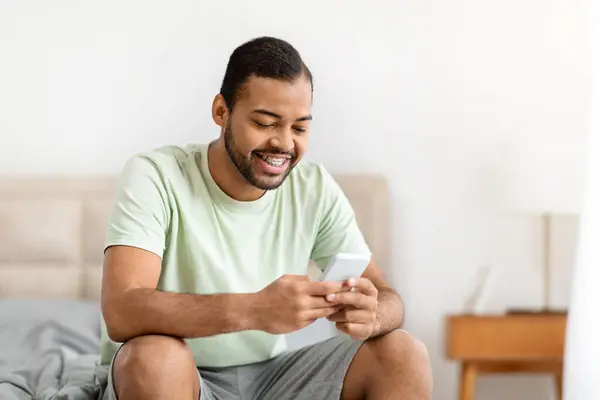 Pozitivní Pohledný Mladý Černoch Pyžamu Sedí Doma Posteli Používá Mobilní — Stock fotografie