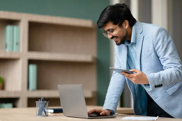 Indian millennial entrepreneur man juggling digital tasks on his laptop and smartphone, stands near office desk indoors, texting and reading emails on computer, managing work and communication