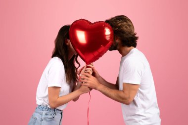 Sevgi dolu beyaz tişörtlü beyaz beyaz bir çift büyük kırmızı bir kalp şeklindeki balonun arkasına gizlenmiş pembe bir arka planda, stüdyoda romantizm ve gizem hissi yaratıyor.